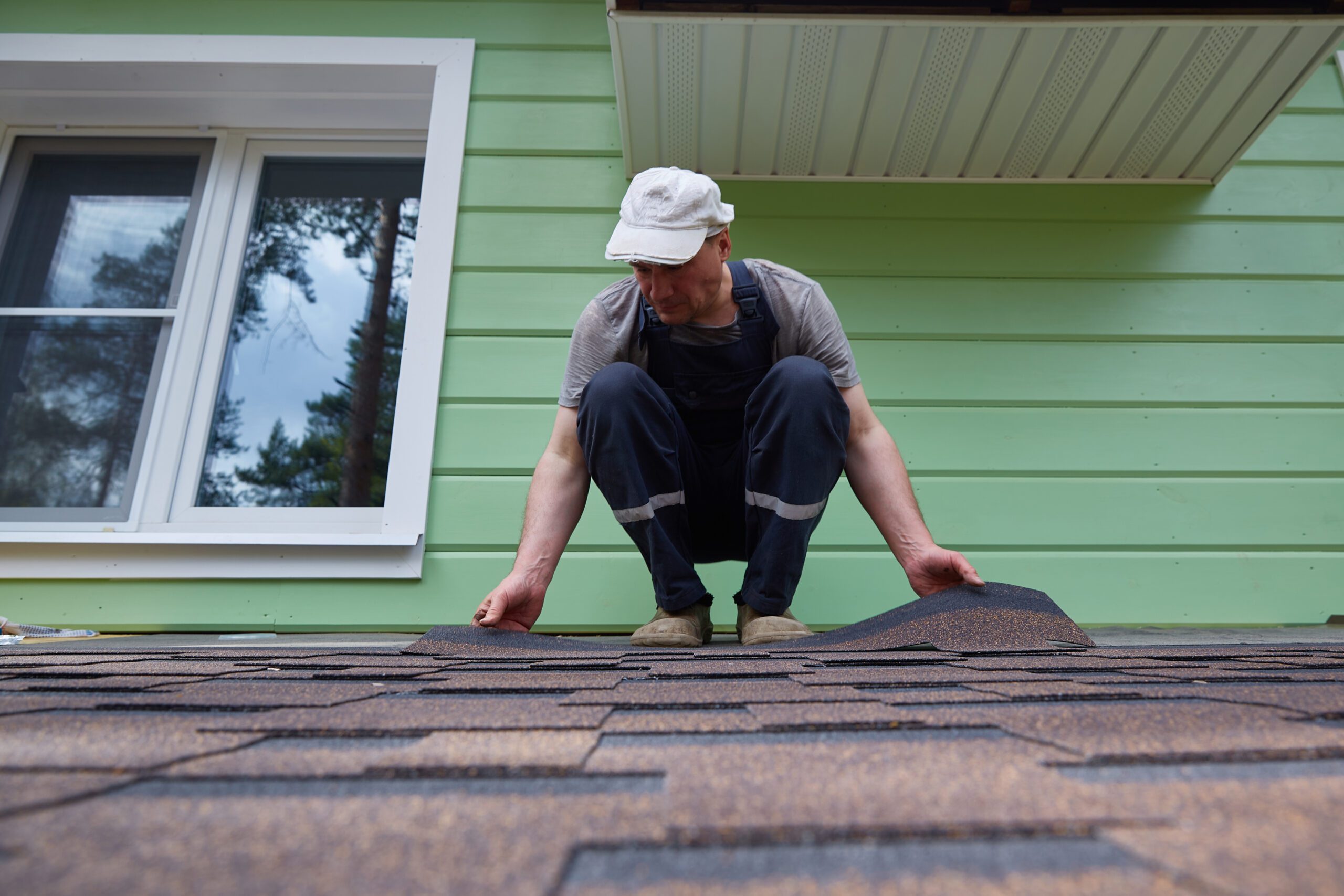 Male worker lays a sheet of soft flexible bitumen shingles on the roof of a country house